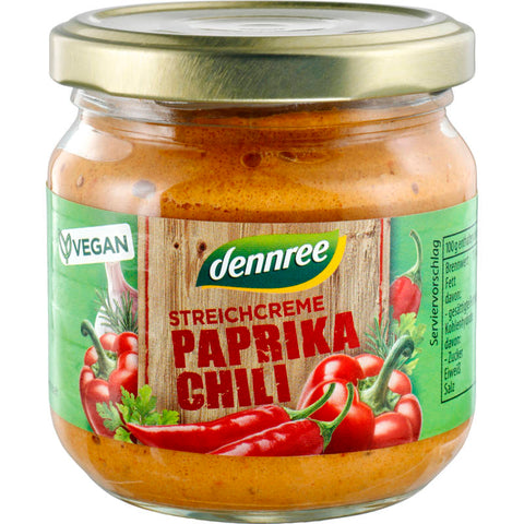 Streichcreme Paprika-Chili in einem Glasbehälter mit rotem Deckel, umgeben von frischen Paprikaschoten und Chilischoten.