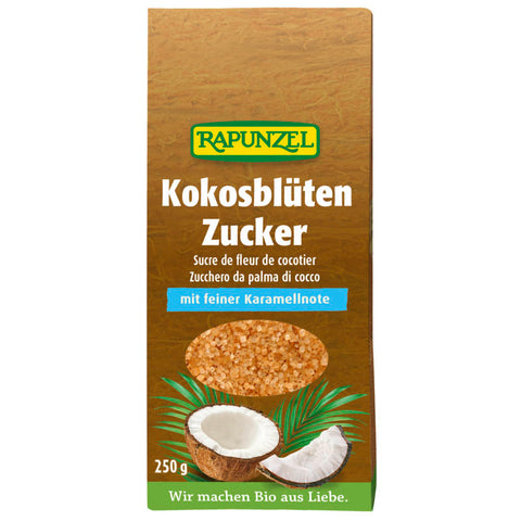 Ein Glasbehälter gefüllt mit braunem Kokosblütenzucker, umgeben von frischen Kokosnüssen und grünen Palmenblättern, auf einem rustikalen Holztisch arrangiert.