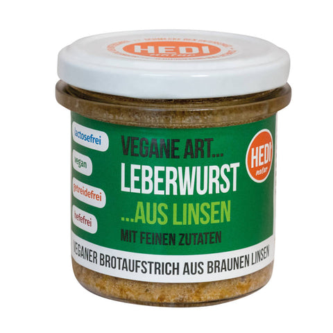 Glas mit veganer Leberwurst aus Linsen, auf einem Holztisch stehend, umgeben von frischen Kräutern und Brot.